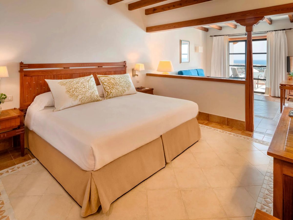A bedroom with a large bed, a carved wooden headboard and a tiled floor, opening past a low partition to a sitting area and a terrace with the sea beyond.
