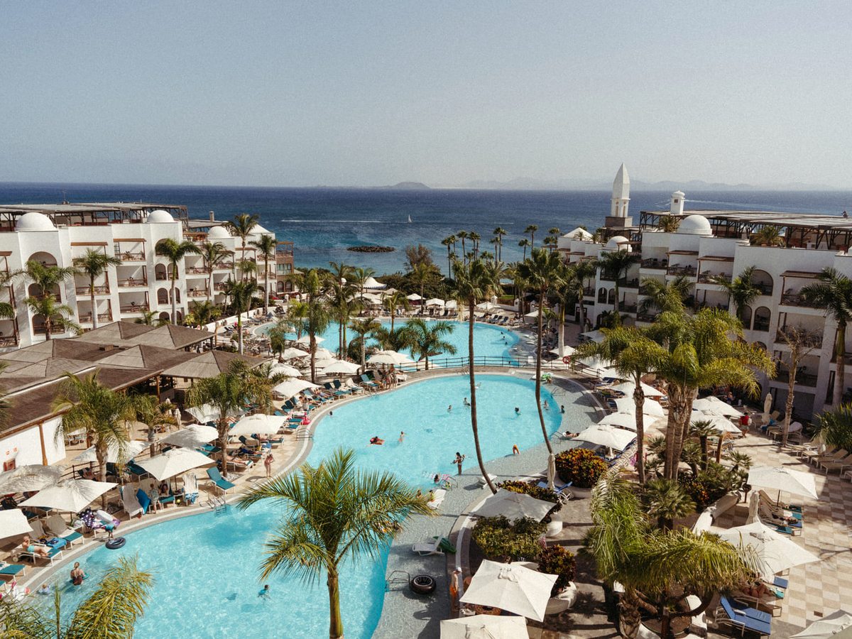 Elevated view of a large lagoon-shaped pool ringed by palms, parasols and sun loungers, with four-storey white resort buildings on either side and the sea behind.