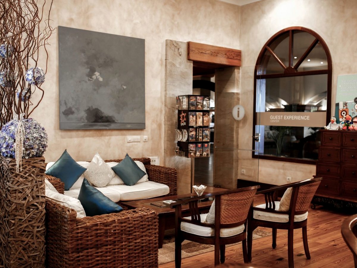 A lounge of wicker sofas with blue cushions and wooden armchairs around low tables, beside a glazed partition signed Guest Experience Center and an arched interior window.