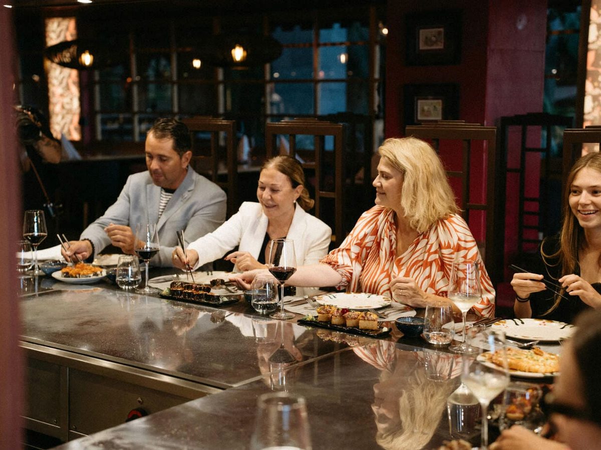 Four diners seated along a polished steel teppanyaki counter with plates of grilled food and glasses of red and white wine, in a warmly lit room with dark wood and red panelling.