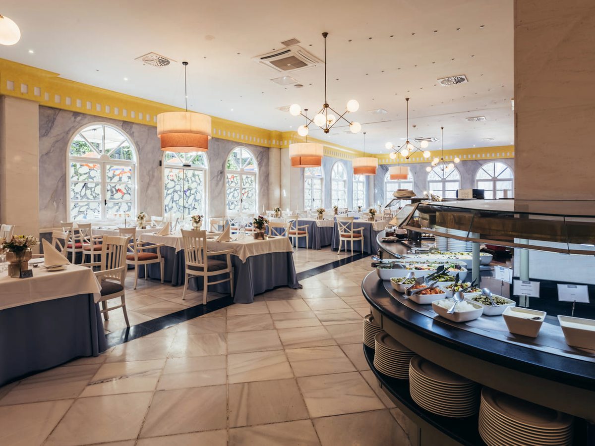 A bright buffet dining room with white-clothed tables and pale wooden chairs beside tall arched windows, and a curved serving counter of salads and cold dishes with stacked plates.