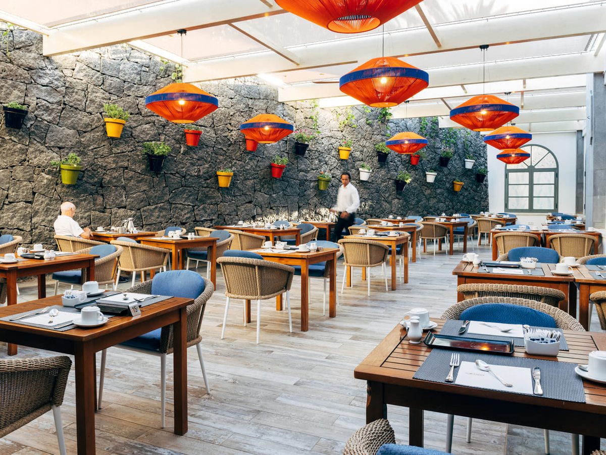A covered terrace dining room with wooden tables laid with place settings, wicker and blue chairs, orange pendant lampshades and a dark volcanic stone wall hung with painted plant pots.