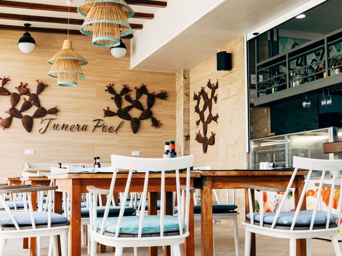 A restaurant interior with white chairs and pale blue cushions at long wooden tables, woven pendant lampshades and a wall of cactus sculptures beside a sign reading Tunera Pool.