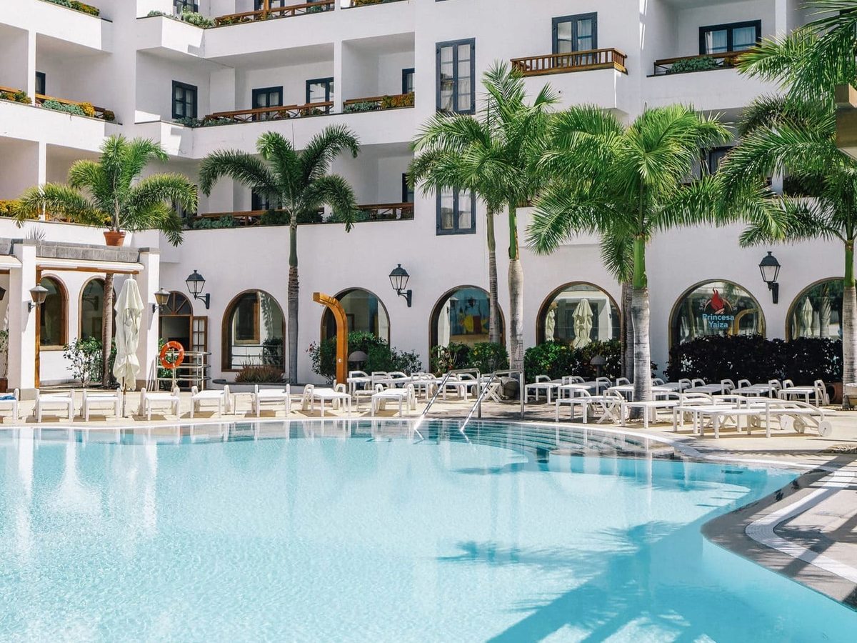 A rectangular pool with white sun loungers along one side, enclosed by a white building with arched ground-floor openings and balconies, with young palms around the edge.