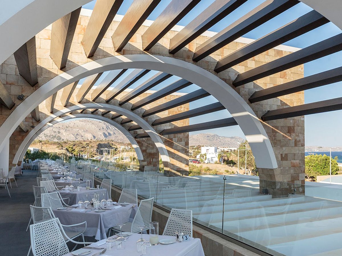 Long open-air dining terrace under stone arches and timber beams, set with white linen tables and white metal chairs, with hills and the coast beyond a glass balustrade.