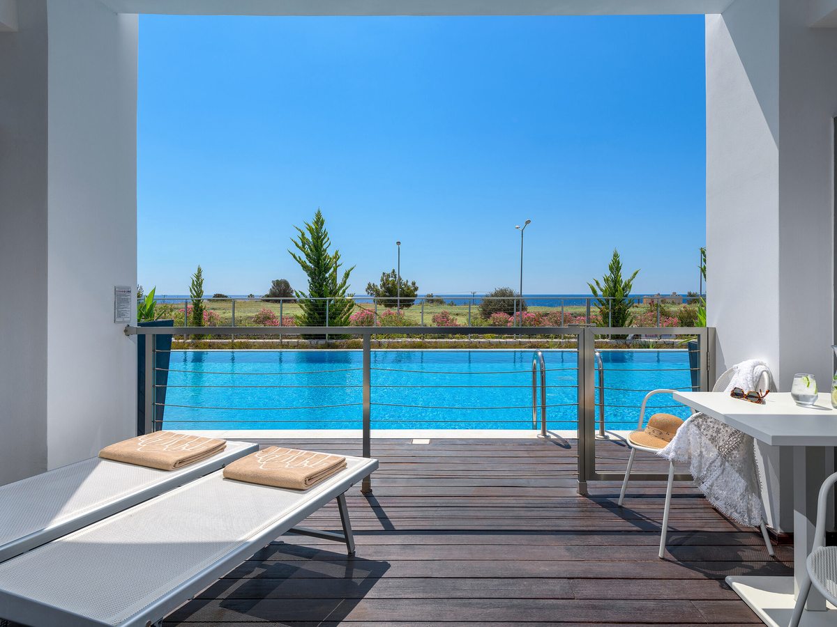 Decked room terrace with two sun loungers and a table and chairs, opening onto a long shared pool with shrubs and the sea beyond.