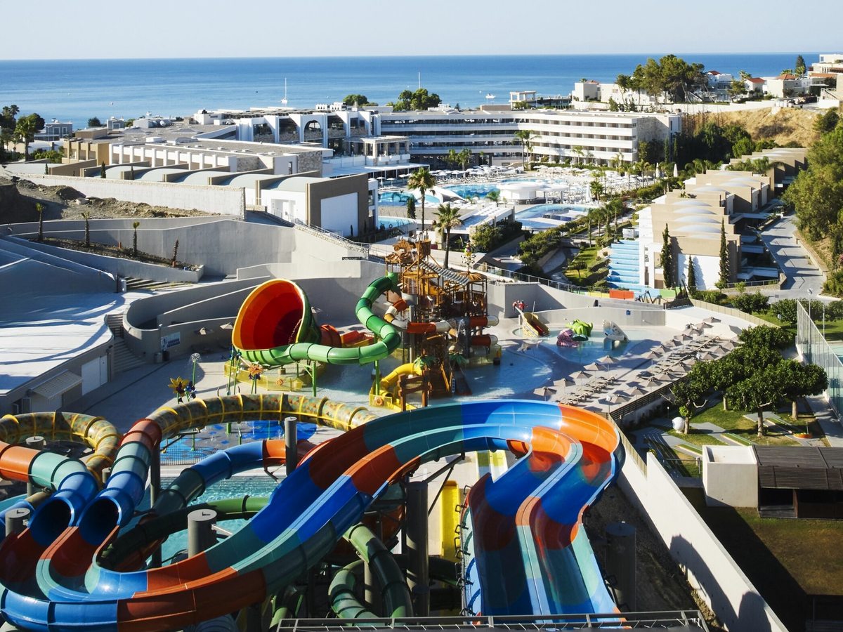 Elevated view over the resort's waterpark, with large multicoloured slide towers in the foreground, low buildings, pools and the sea behind.