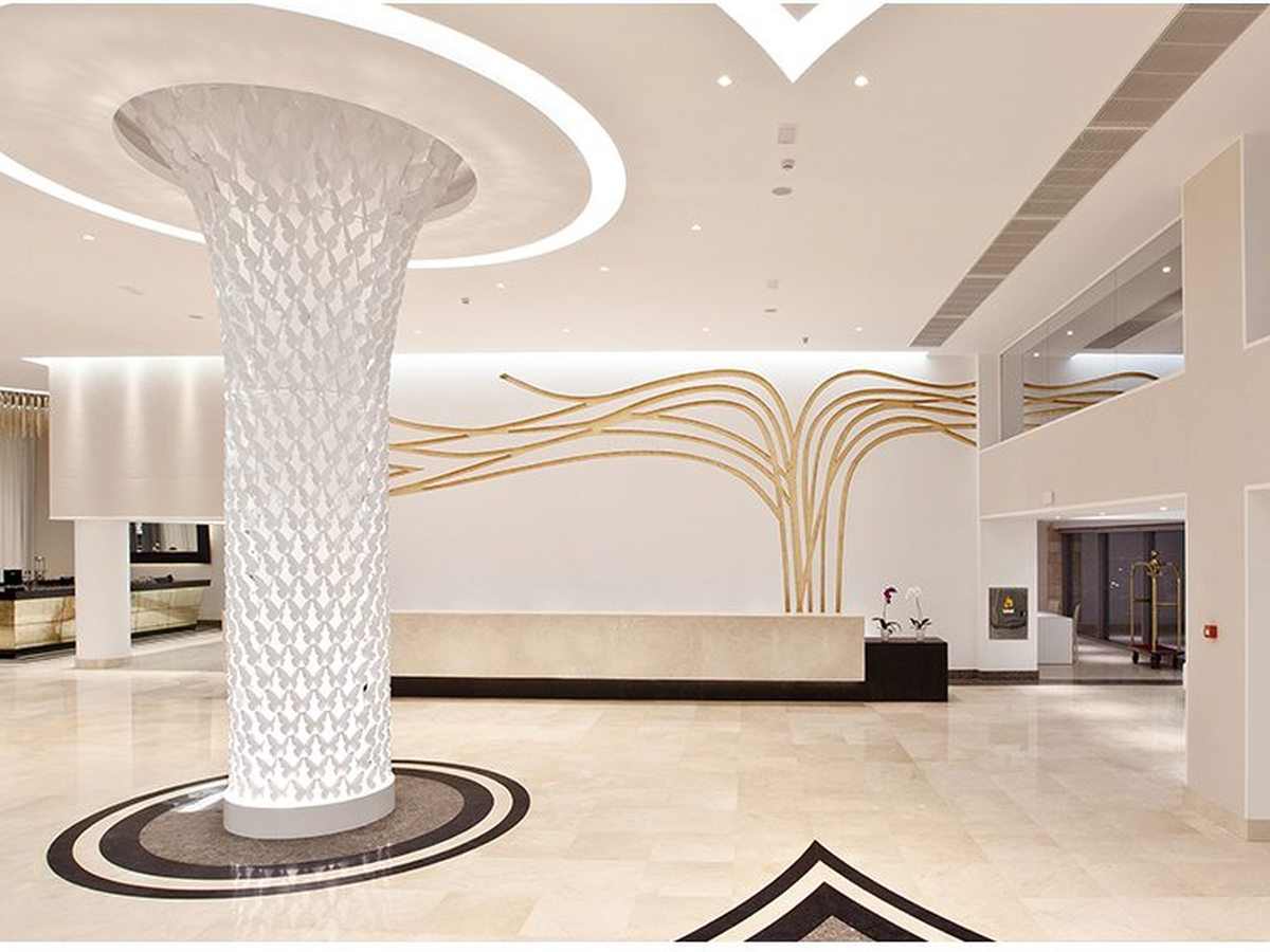 Marble-floored hotel lobby with a tall white sculptural column, a low stone reception desk and a gold branch design across the wall behind it.