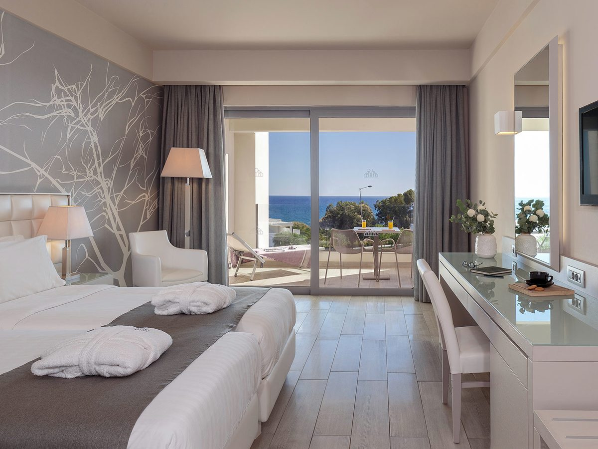 Twin-bedded room with a grey branch-patterned feature wall, a white glass-topped desk and sliding doors onto a balcony looking out to the sea.