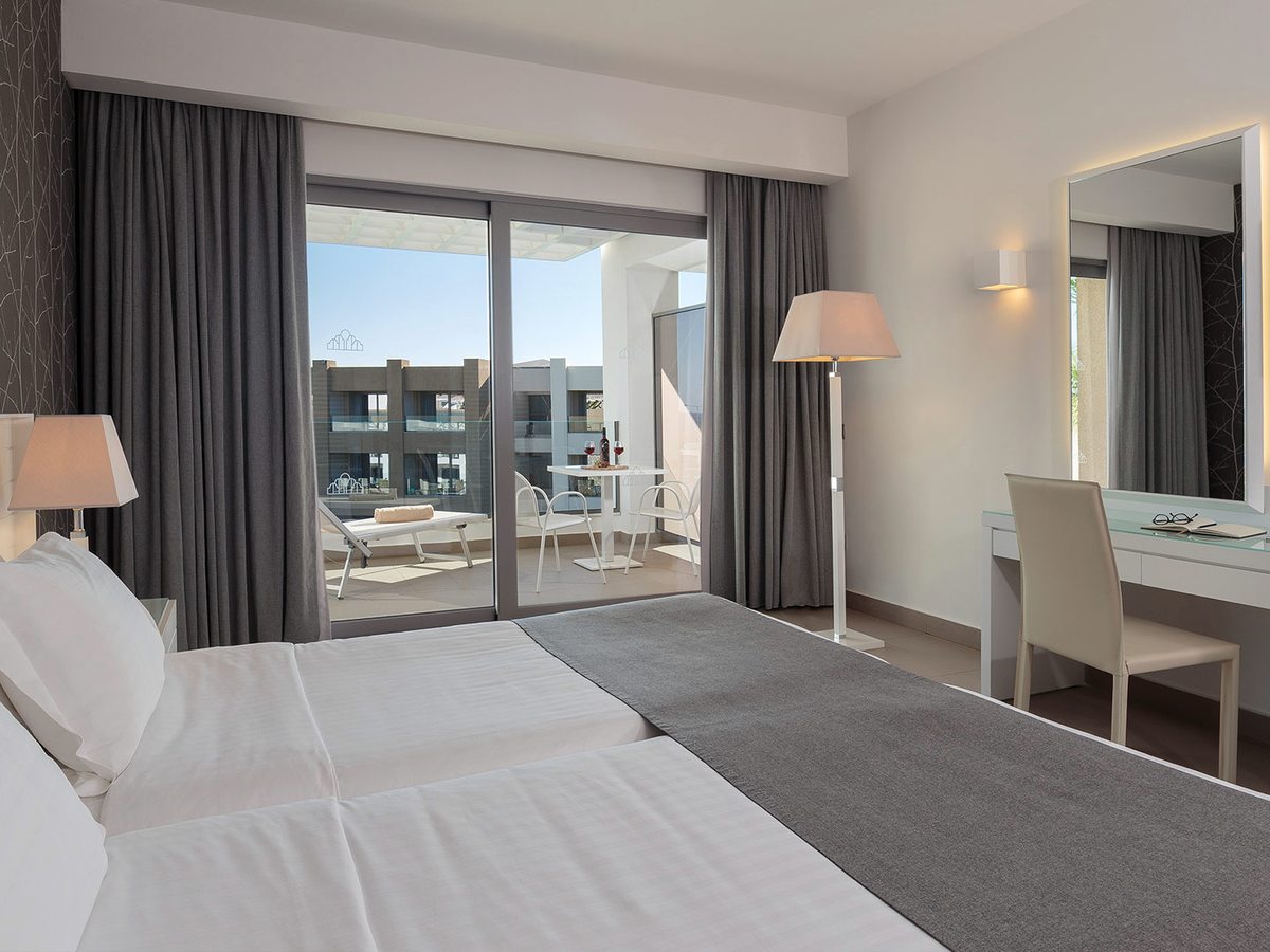 Family room with a large bed, a floor lamp and a white dressing table, and sliding doors onto a balcony with a table, chairs and a sun lounger.