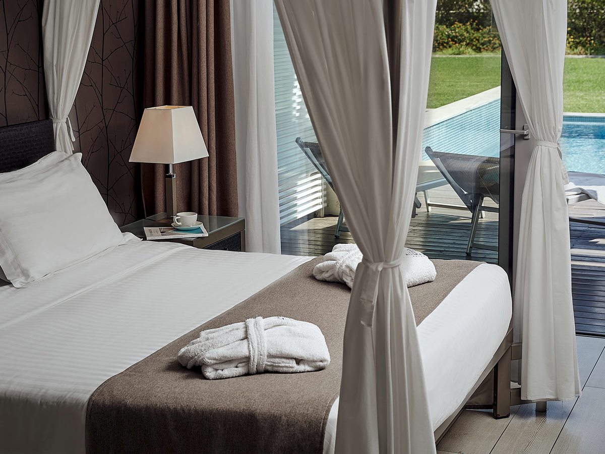 Four-poster bed dressed in white and brown linen, with glass doors open to a decked terrace, sun loungers and a private pool on a lawn.