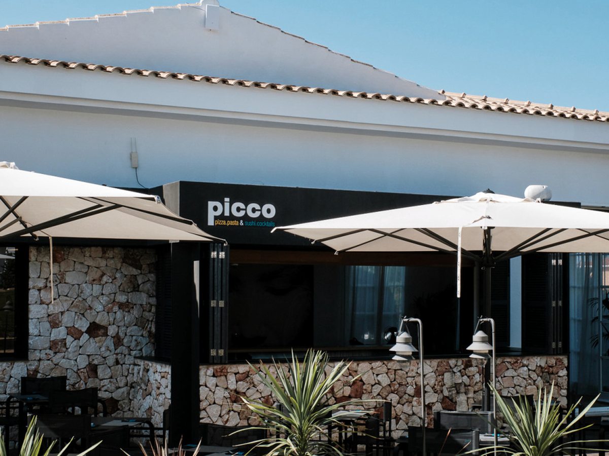 The whitewashed and stone-clad exterior of Picco restaurant, its black sign reading picco, pizza, pasta and sushi, cocktails, with cream parasols over patio tables.
