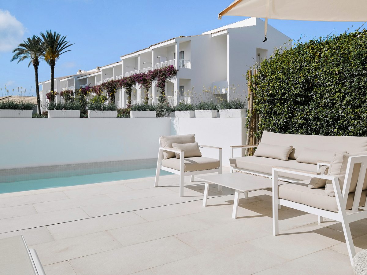 A narrow private pool on a paved terrace with white-framed sofas and armchairs under a parasol, screened by a white wall and hedge, with a guest-room block beyond.