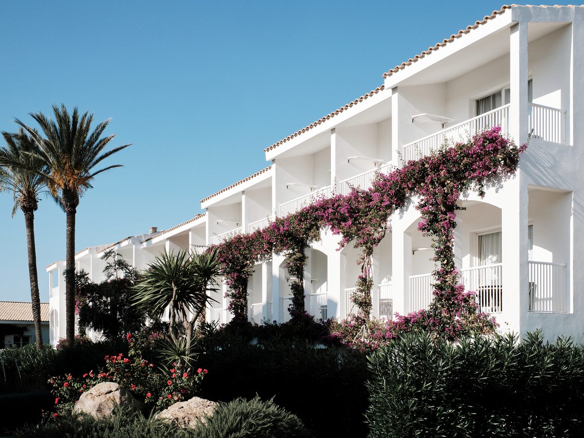 A long white two-storey accommodation block with white balcony railings, draped with pink bougainvillea, with palms and planted borders in front under a clear blue sky.