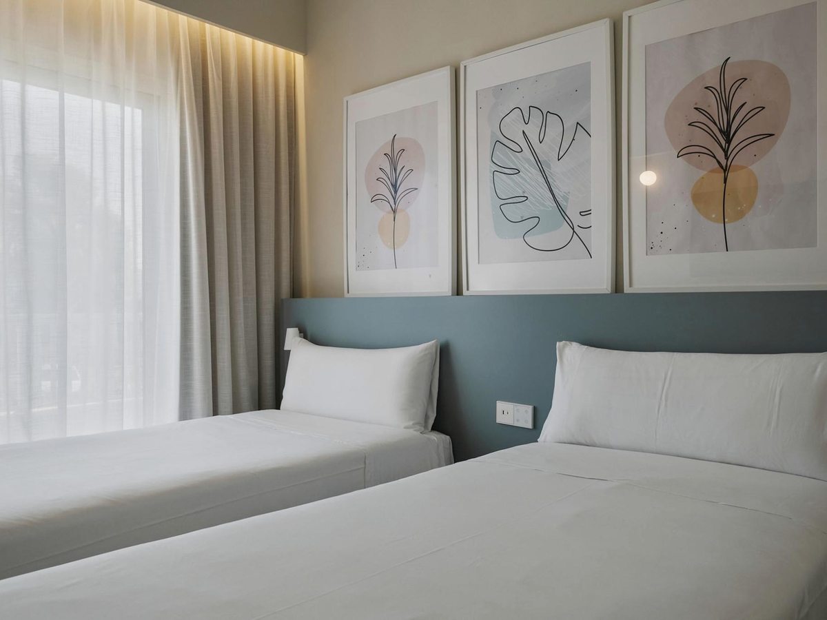 A guest room with two single beds against a teal upholstered headboard, three framed botanical prints above and a full-height window with sheer curtains.