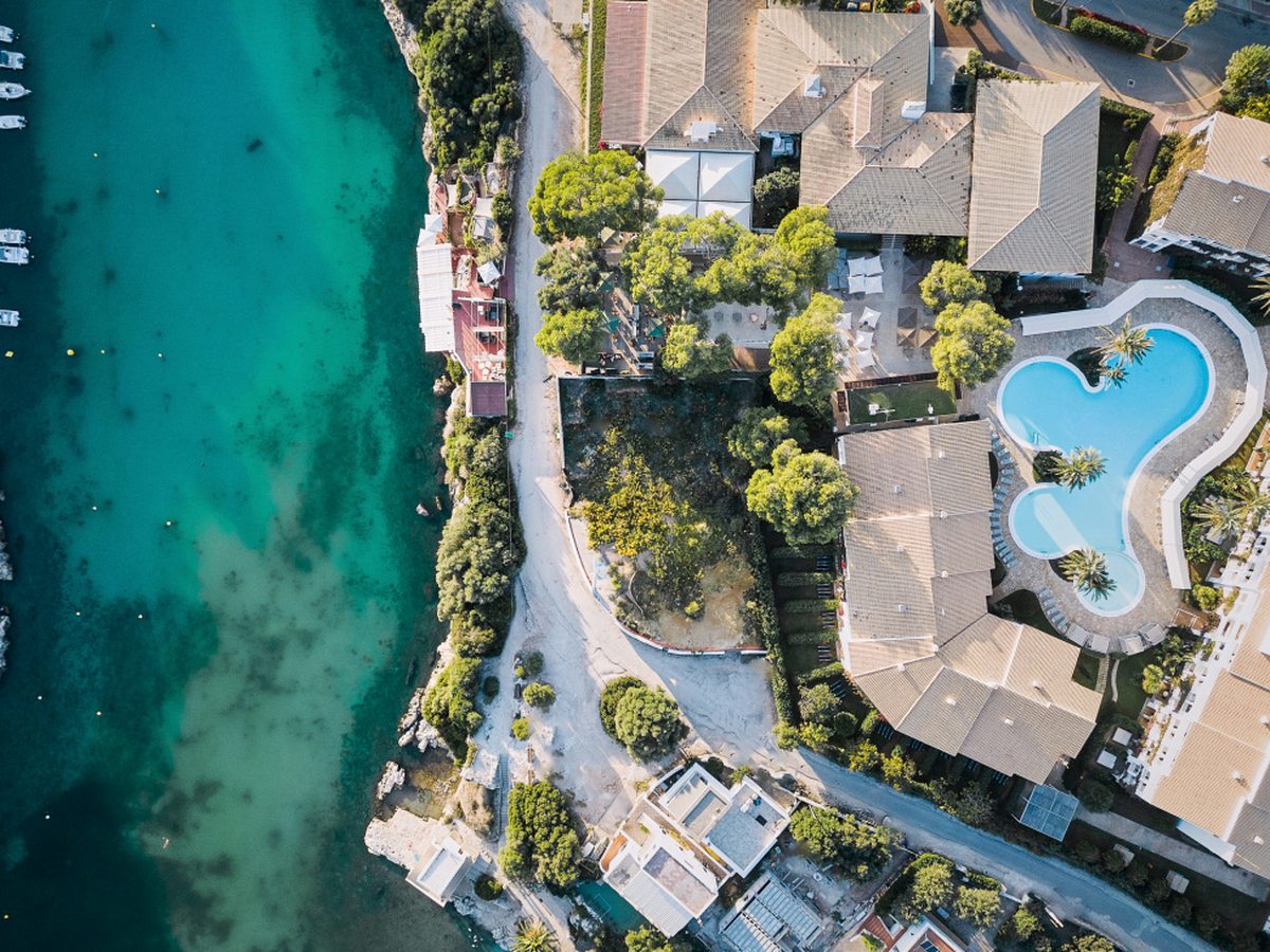 Overhead aerial of the hotel's tiled roofs, gardens and freeform pool on one side of a narrow turquoise inlet, with small boats moored along the far shore.