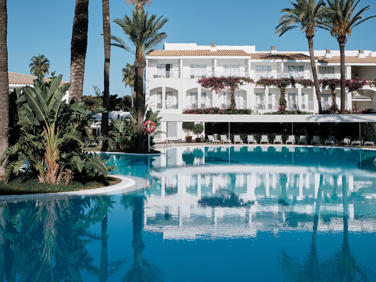 A large freeform pool reflecting a white two-storey accommodation block draped with bougainvillea, with sun loungers along a shaded terrace and tall palms and banana plants around it.