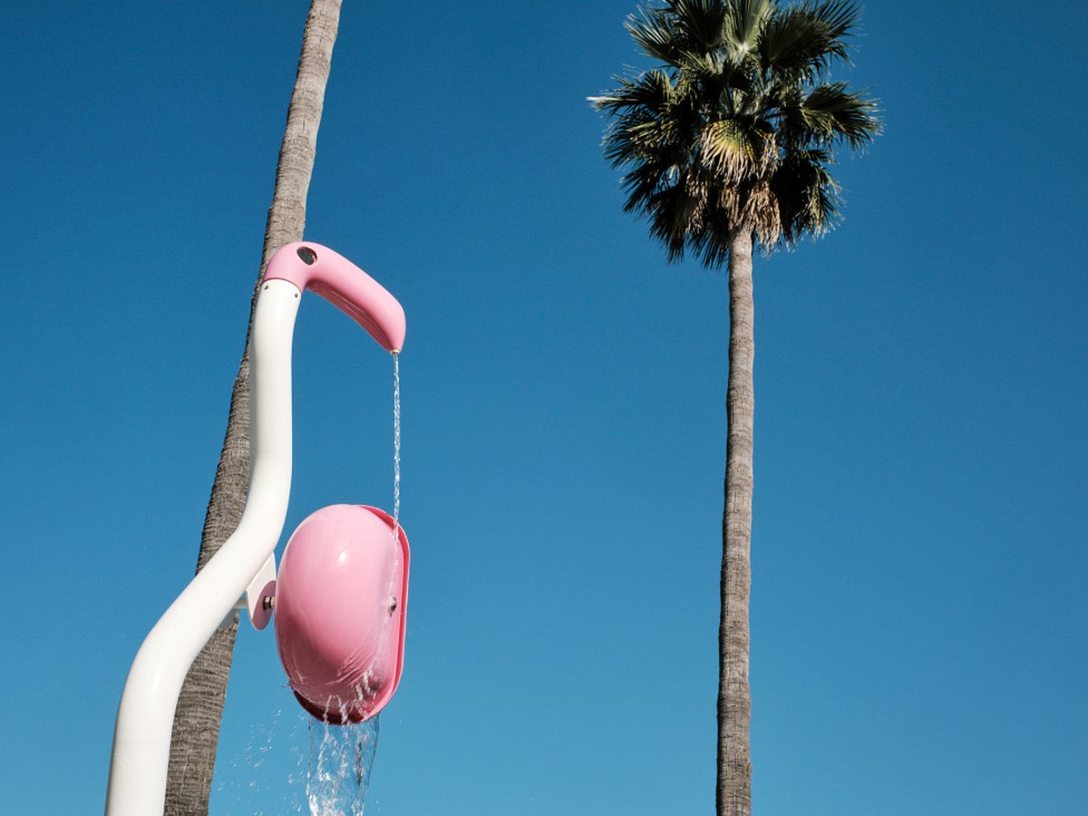 A pink and white flamingo-shaped tipping water bucket pouring water, against a deep blue sky with two palm trees behind.
