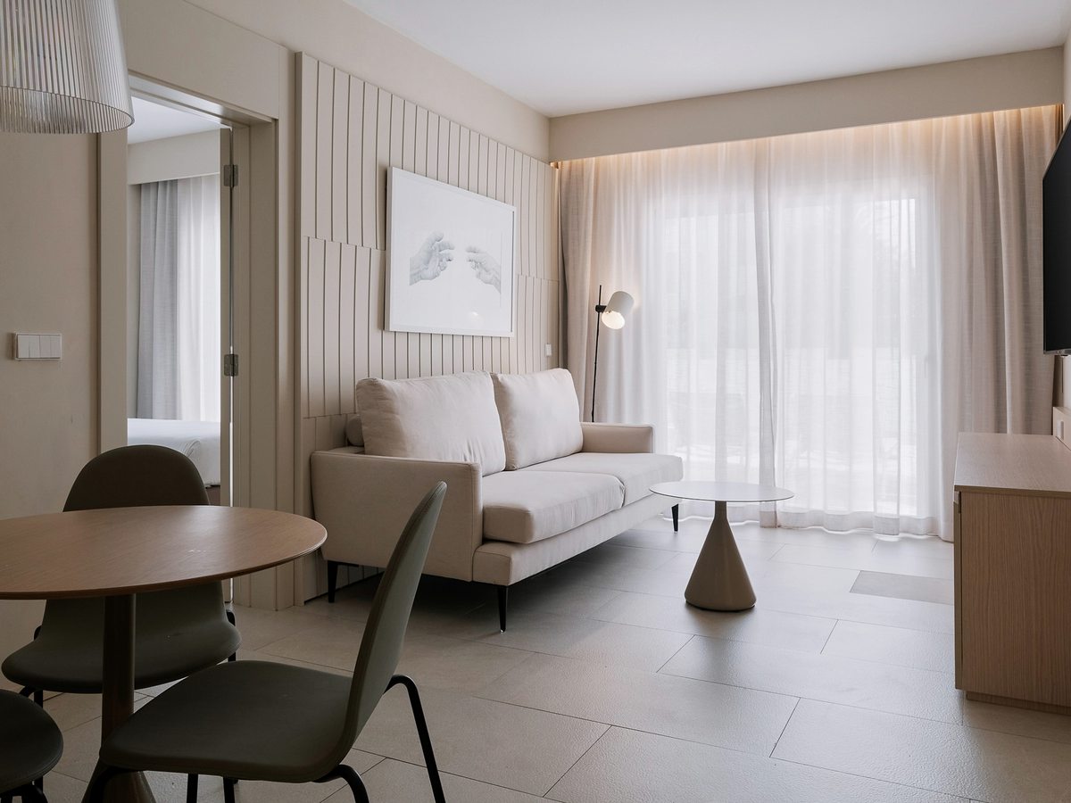 A suite living area with a cream sofa, a round wooden dining table and chairs, a wall-mounted television and full-height windows with sheer curtains, and the bedroom through an open door.