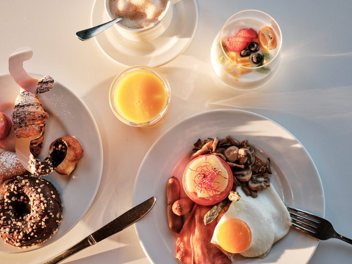 A breakfast table with fried eggs, bacon, mushrooms and tomato, pastries and a doughnut, pots of fruit and yoghurt, orange juice and coffee.