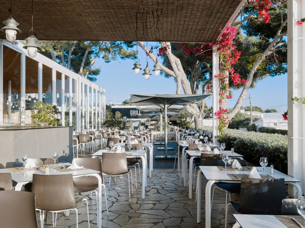 A covered restaurant terrace with white tables set with wine glasses and napkins, cushioned chairs, hanging lanterns and pink bougainvillea, with the black GRILL sign beyond.
