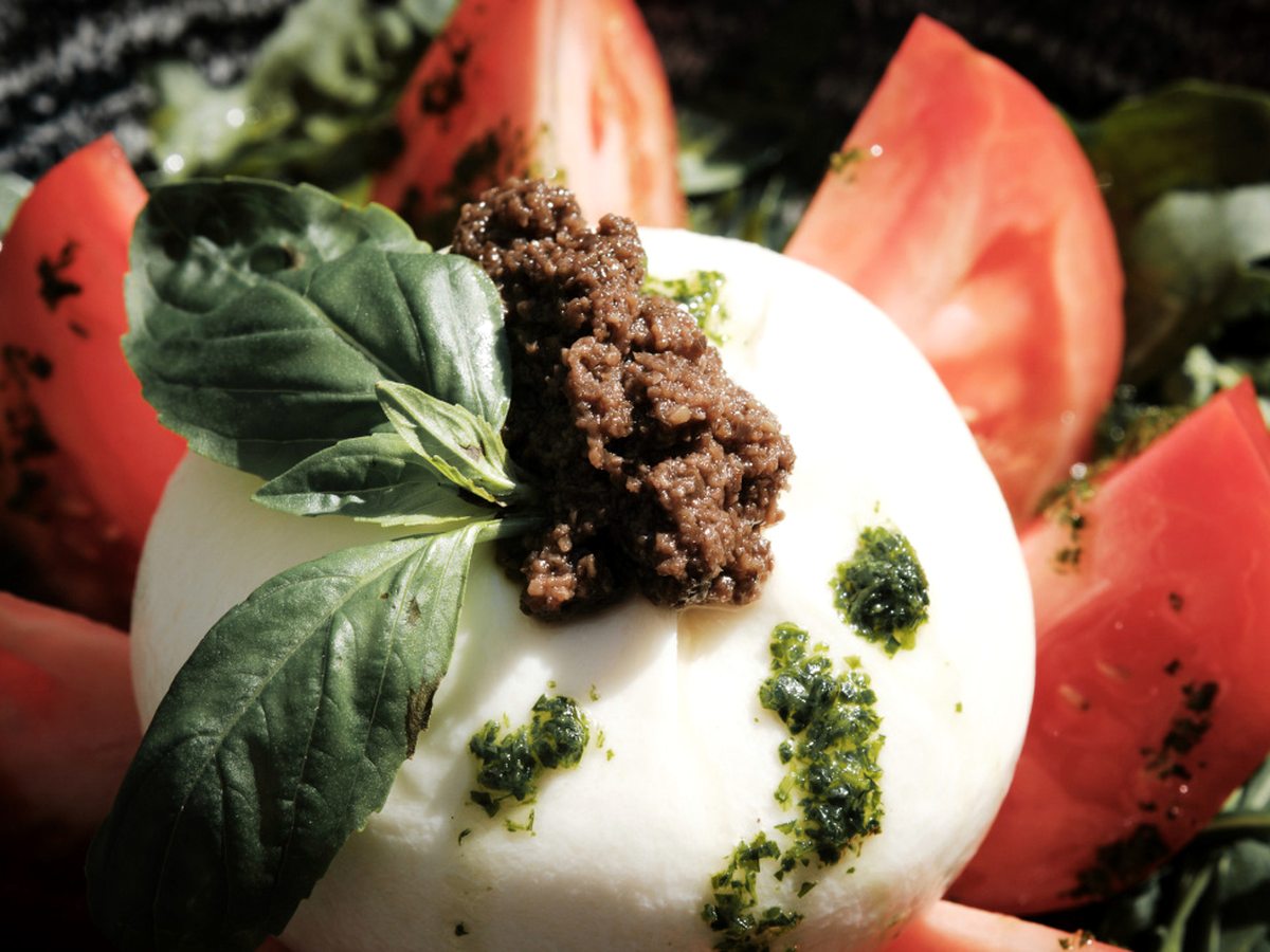 A plate of burrata with tomato wedges, fresh basil leaves, olive tapenade and basil oil.