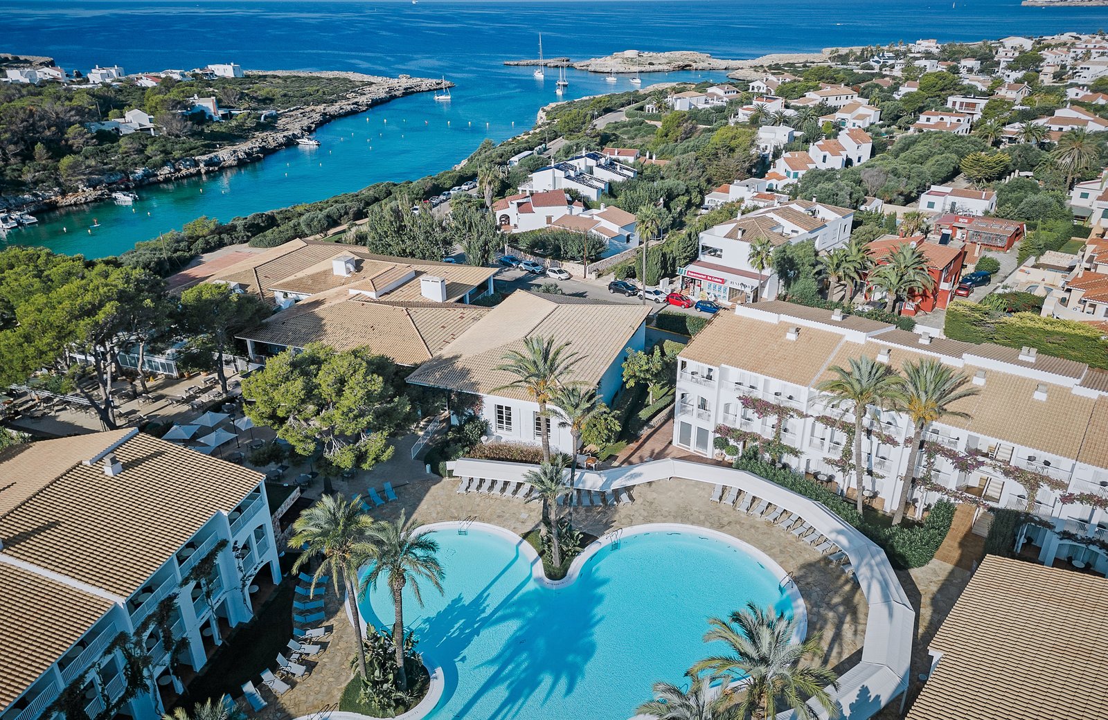 Aerial view of Prinsotel La Caleta: a freeform pool ringed by sun loungers and palms, with low white buildings under tiled roofs around it and a turquoise coastal inlet and the sea beyond.
