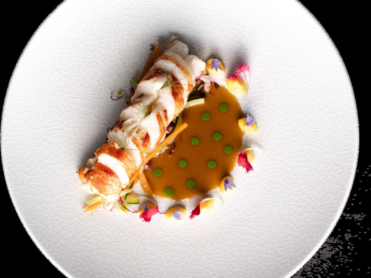 A plated fine-dining dish of lobster medallions in an orange sauce with dots of herb oil, small vegetables and edible flowers on a white textured plate.