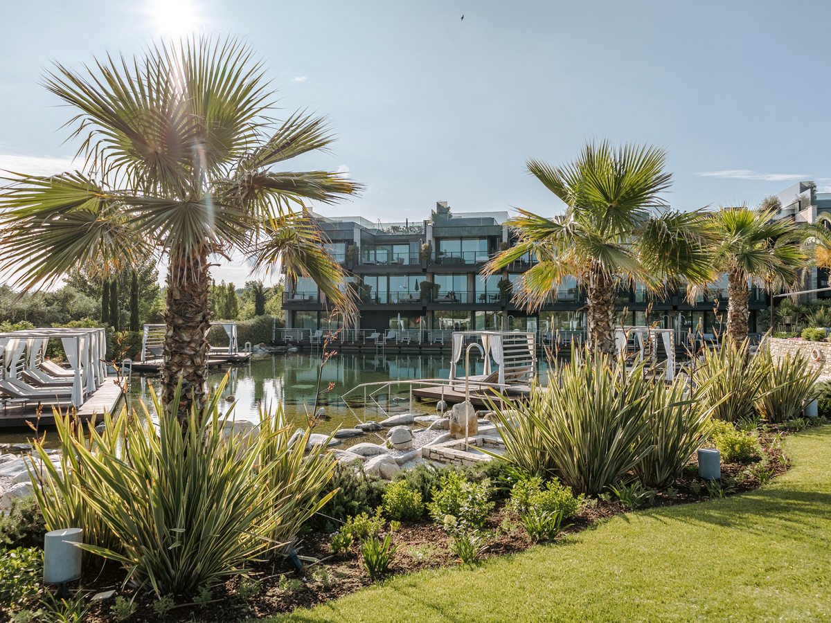 A landscaped bathing pond with rock and gravel edges, timber decks and cabanas, fan palms and grasses in the foreground and the glass-fronted accommodation block reflected in the water.