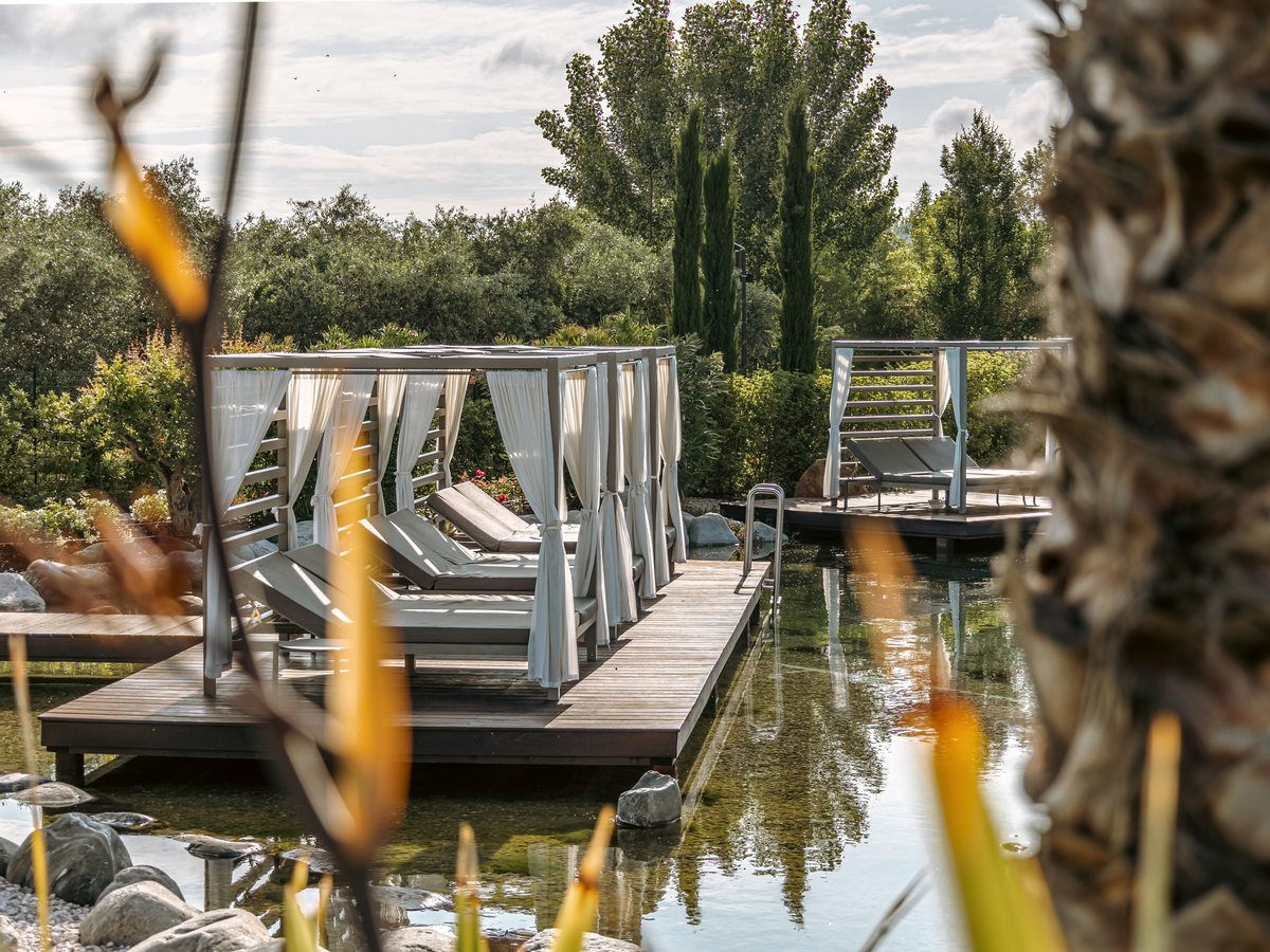 Timber platforms built out over the water carrying curtained cabanas and grey sun loungers, with rocks and reeds at the water's edge and cypress and olive trees behind.