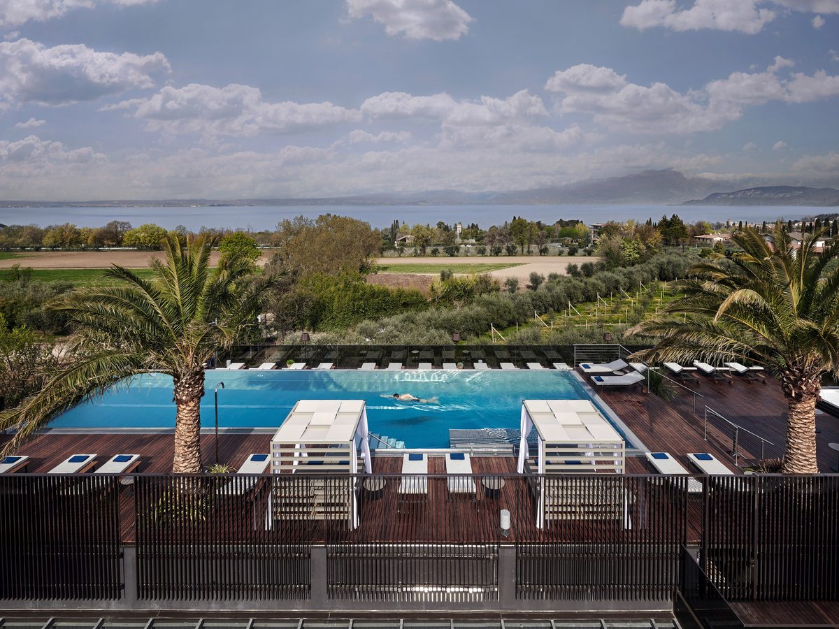 A rectangular rooftop pool on a timber deck with one swimmer doing lengths, canopied double daybeds and palms around it, and Lake Garda beyond the olive groves.