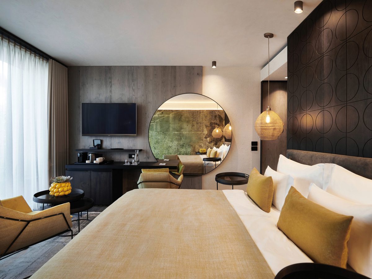 A double room with a mustard throw and cushions on a king-size bed, a dark patterned headboard wall, a round illuminated mirror, a drinks station and a wall-mounted television.