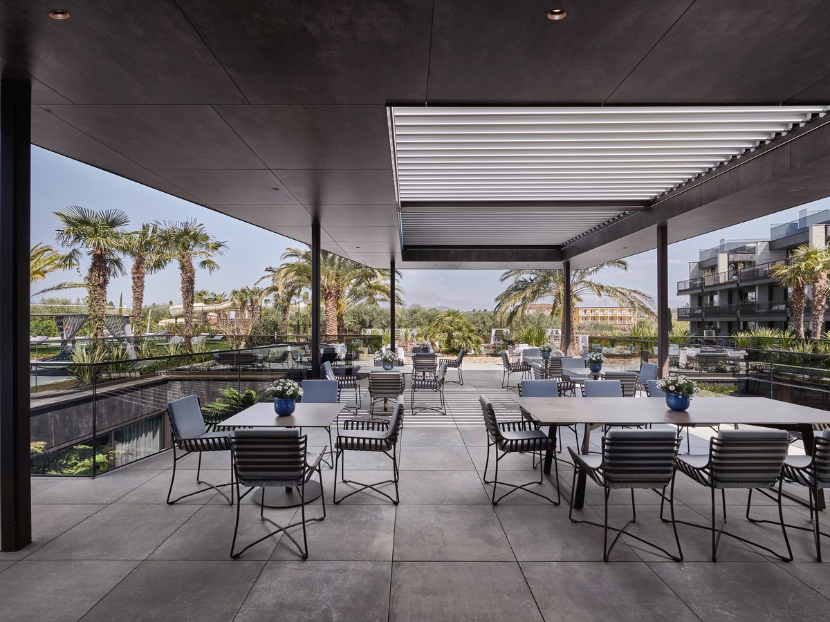 A covered outdoor dining terrace with grey tables and metal-framed chairs under a louvred canopy, with palms, gardens and the pools visible below the glass balustrade.