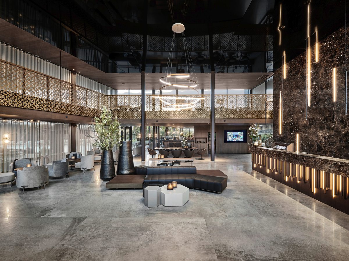 A hotel lobby with a long dark marble reception desk lit from within, black leather bench seating and grey armchairs on a polished stone floor, beneath a gold-screened mezzanine gallery.