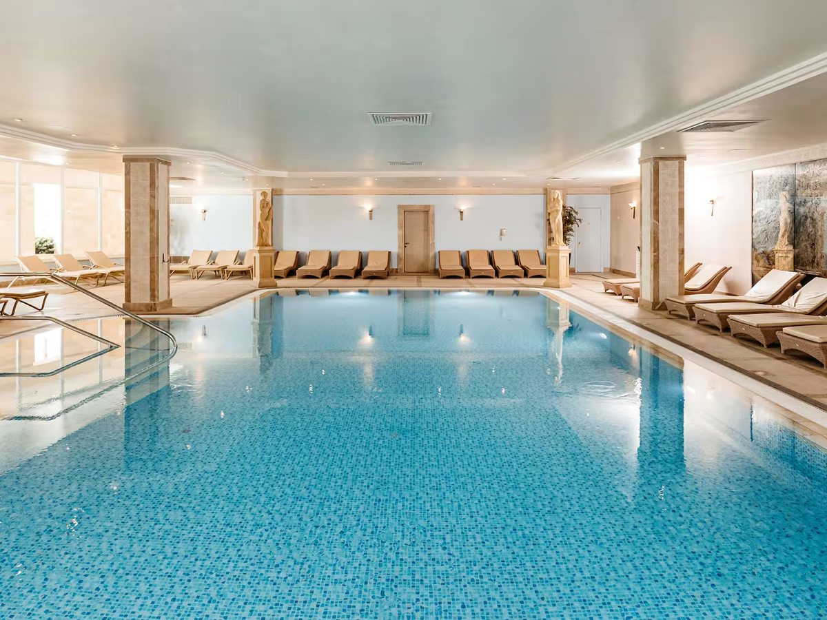 Large indoor pool with pale stone columns, cushioned loungers along both sides and daylight from tall windows.