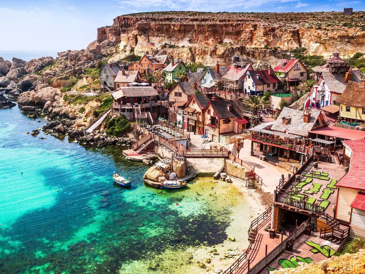 Cluster of brightly painted timber buildings on a rocky cove, with turquoise sea and small boats below.