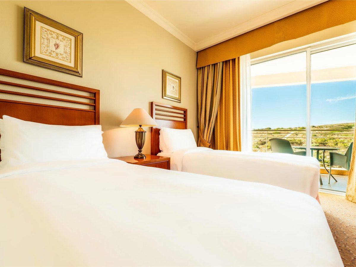 Twin room with two single beds, wooden headboards, framed prints and sliding doors onto a balcony looking over open countryside.