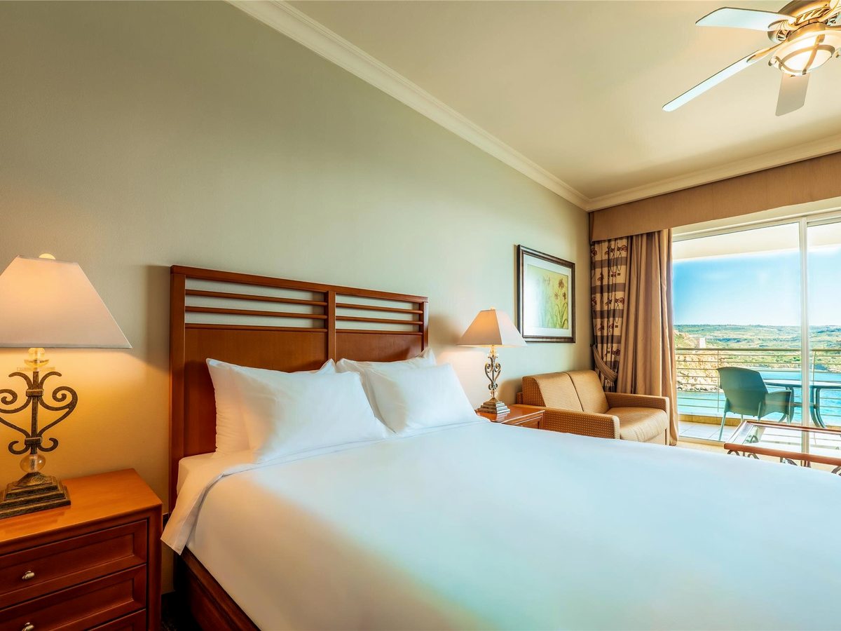 Double room with a slatted wooden headboard, bedside lamps, a wicker armchair and sliding doors onto a balcony with a bay view.
