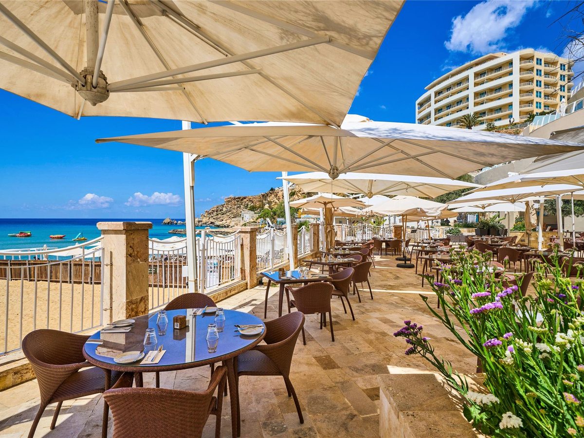 Outdoor restaurant terrace with laid tables, wicker chairs and large cream parasols beside the sand, with the resort tower on the cliff behind.