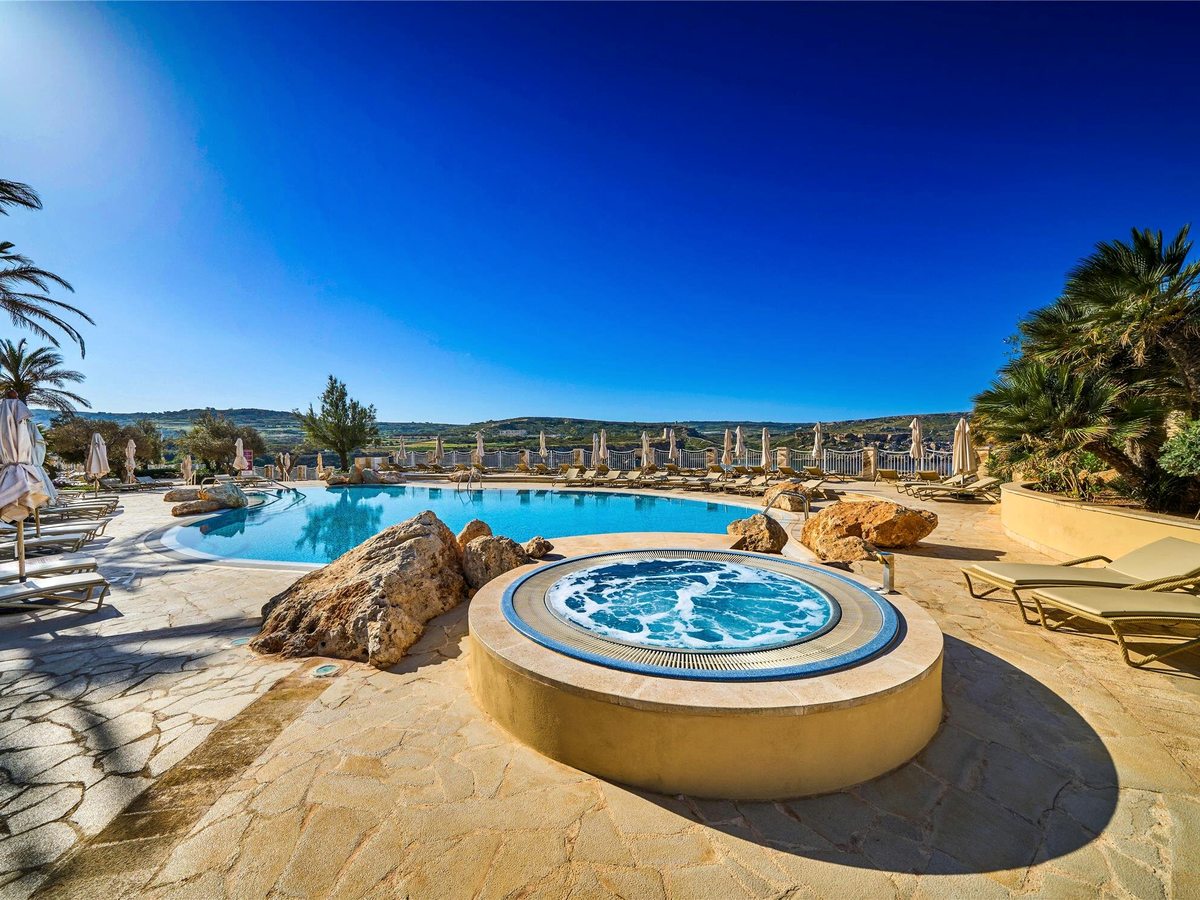 Circular whirlpool raised on a stone plinth on a flagged terrace, with a free-form pool, loungers and parasols behind and low hills in the distance.