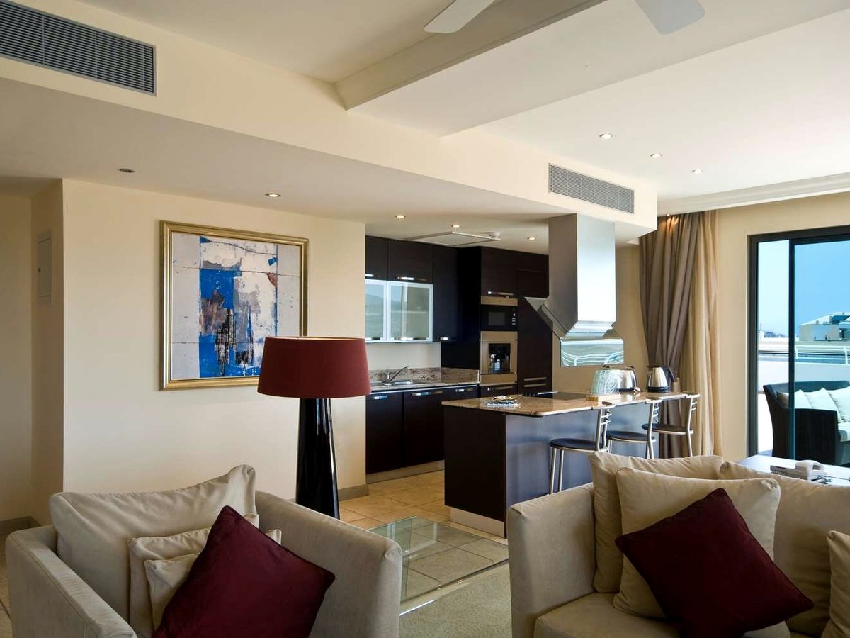 Open-plan suite with cream sofas in the foreground and a dark fitted kitchen with a breakfast bar and stools beyond, next to balcony doors.