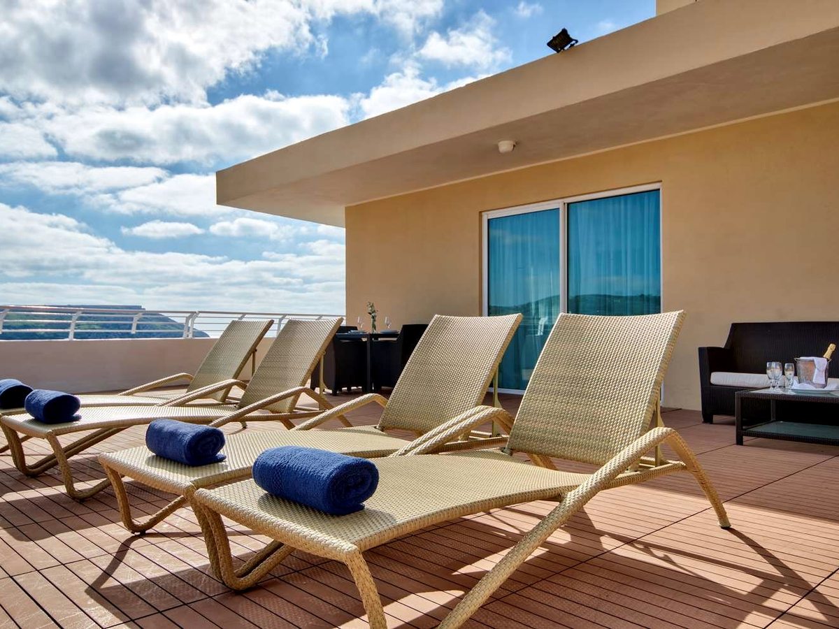 Wide decked terrace with four wicker sun loungers and rolled blue towels, a dining table and a sofa beside the suite's sliding doors.