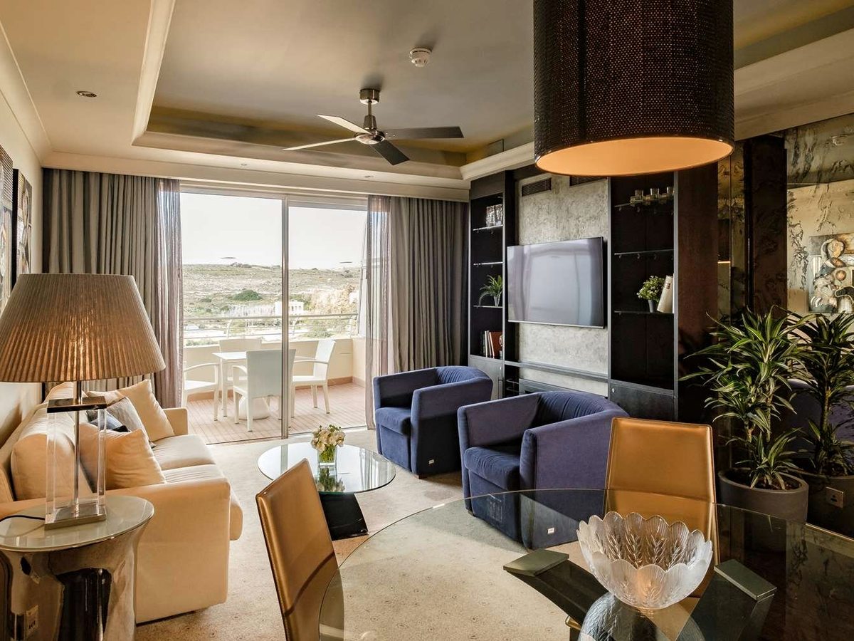 Suite lounge with a cream sofa, two blue armchairs, a glass dining table and a wall-mounted television, opening onto a balcony with a countryside view.