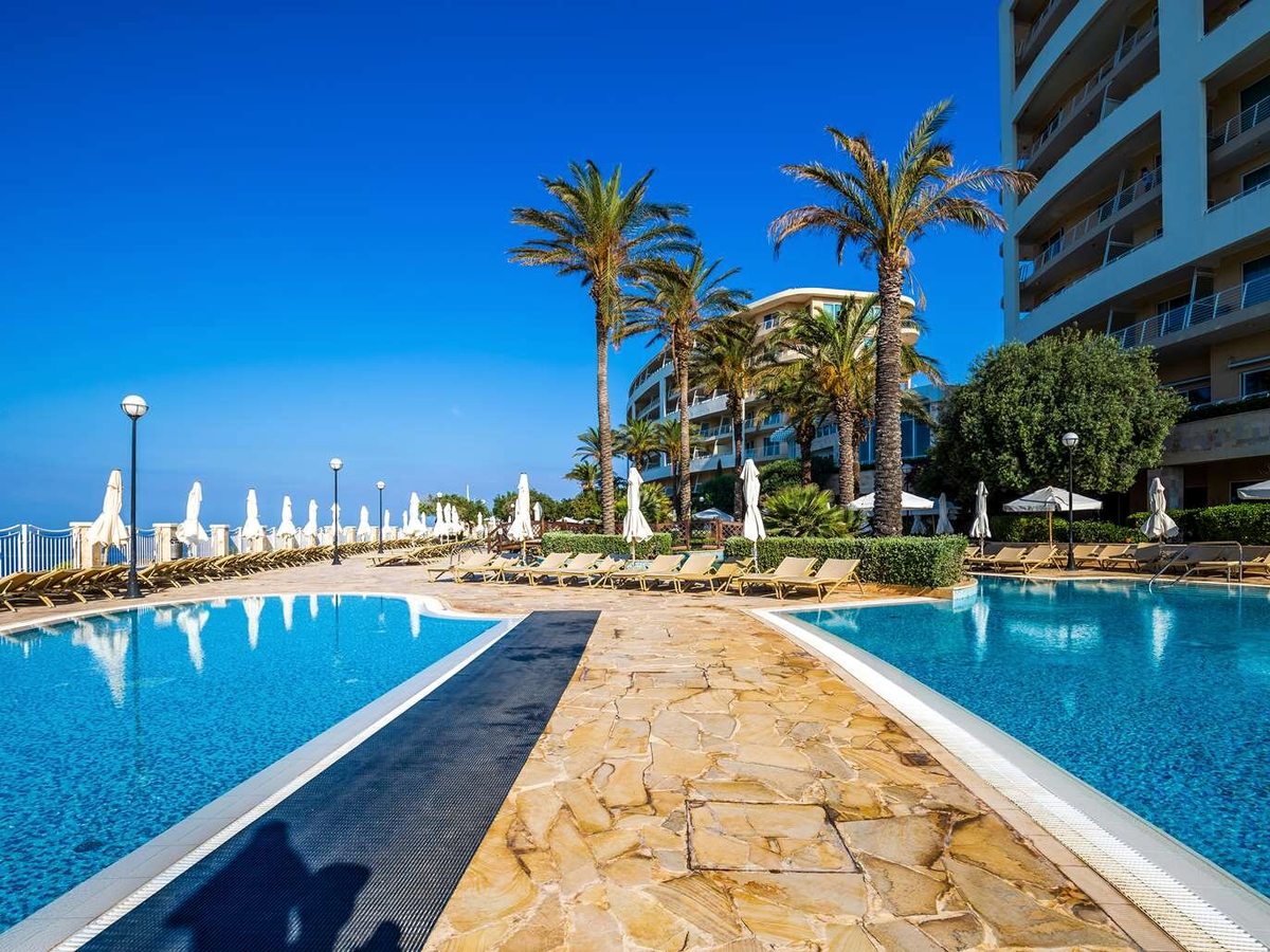 Two outdoor pools either side of a stone walkway, with rows of sun loungers, closed white parasols, tall palms and the resort towers behind.