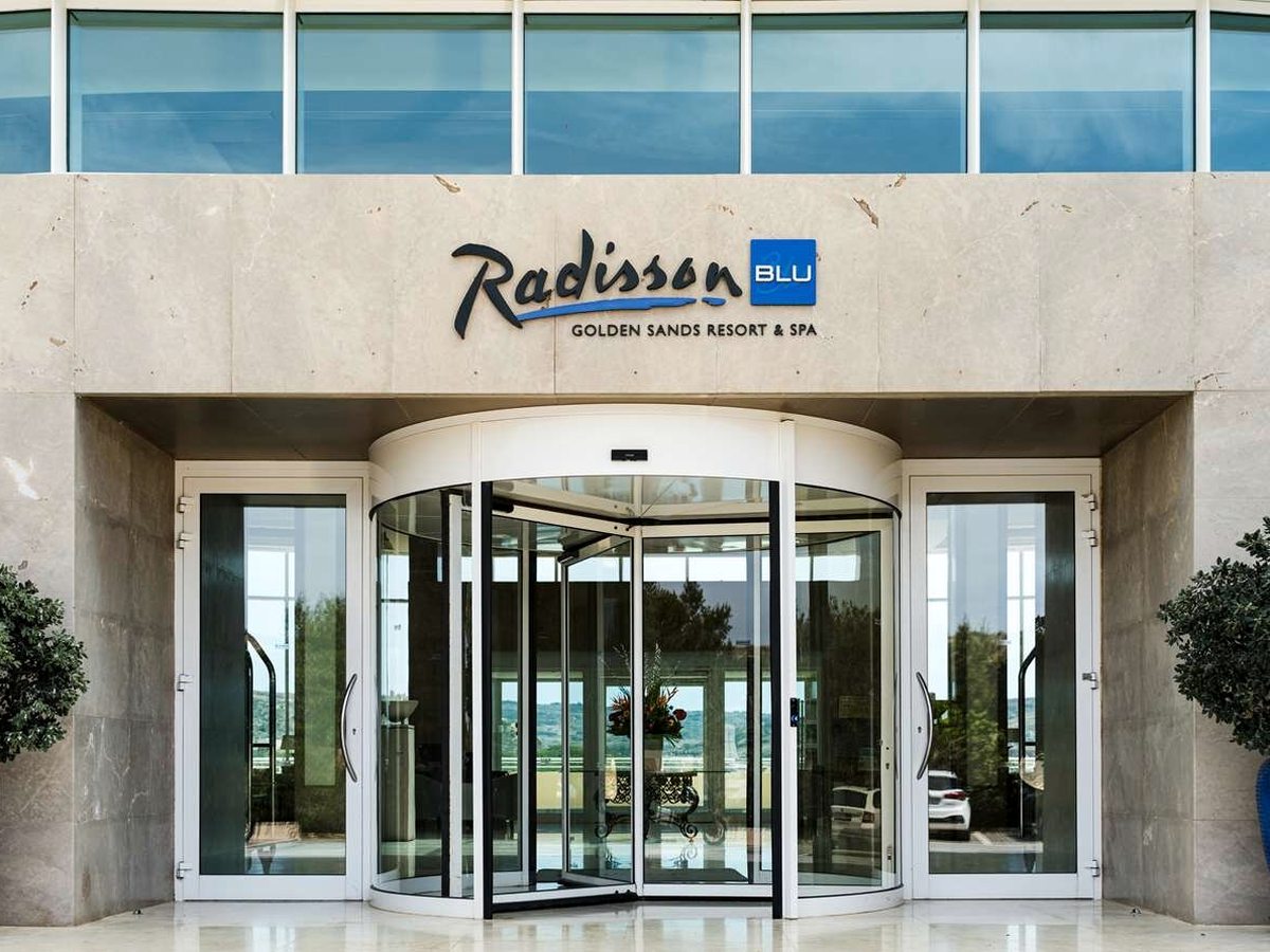 Stone-clad hotel entrance with a revolving glass door and a Radisson Blu Golden Sands Resort and Spa sign above it, flanked by clipped bay trees in planters.