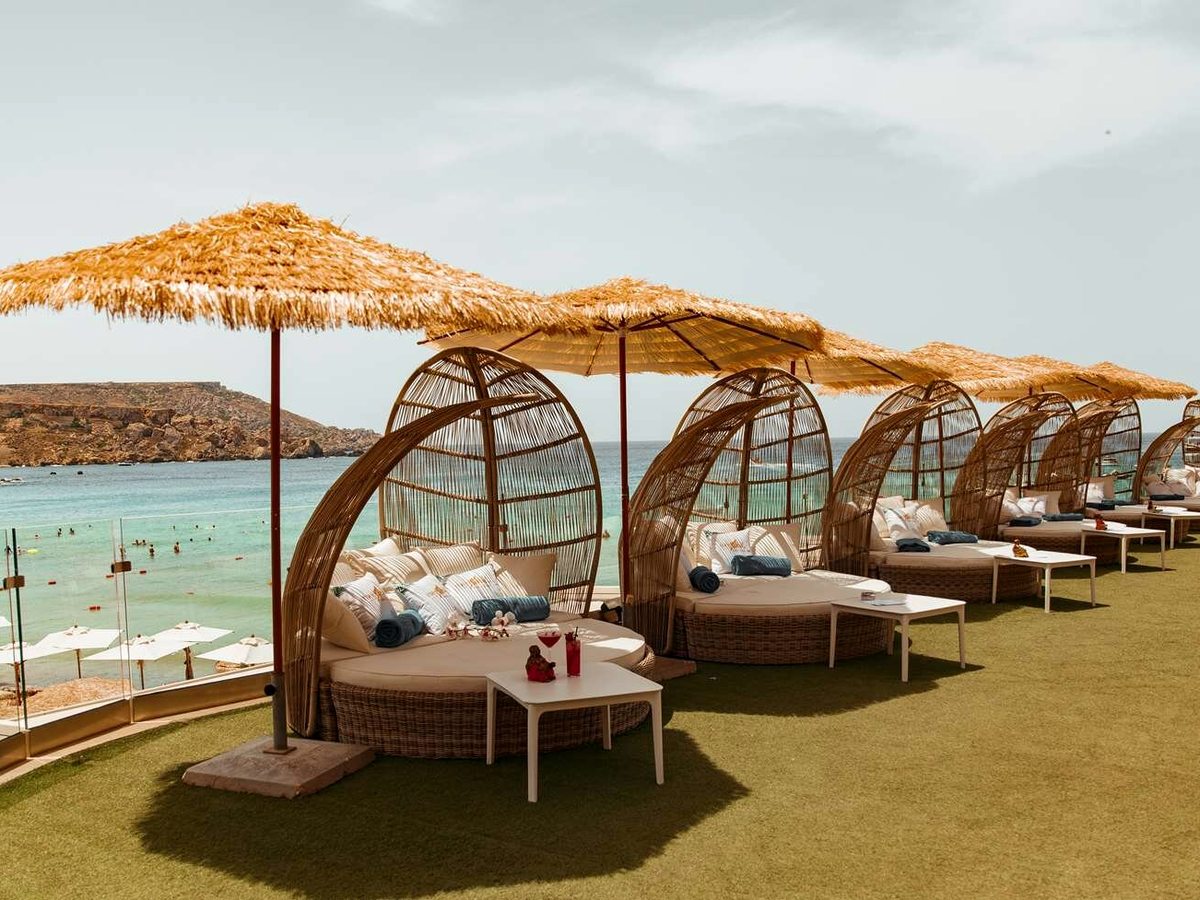 Row of round wicker cabana daybeds with cushions and rolled towels under straw parasols on a lawn terrace, with the bay and the beach below.