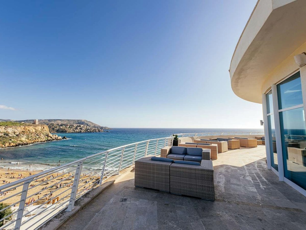 Stone terrace lined with wicker sofas along a white railing, the curved end of the hotel building to the right and Golden Bay's sand and turquoise water below.