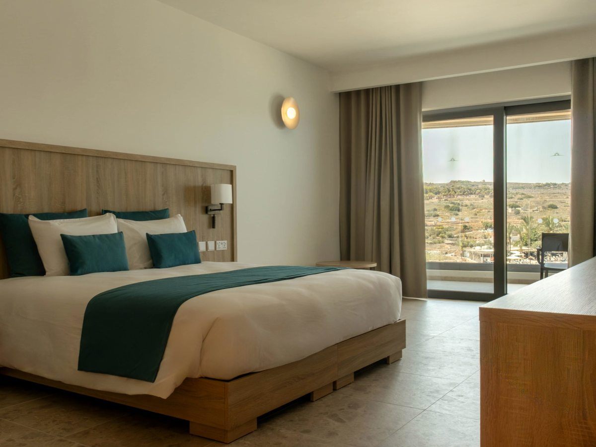 A double bed with a pale wooden headboard and dark green runner, a wall light above the bedside table and full-height balcony doors showing dry open countryside beyond.