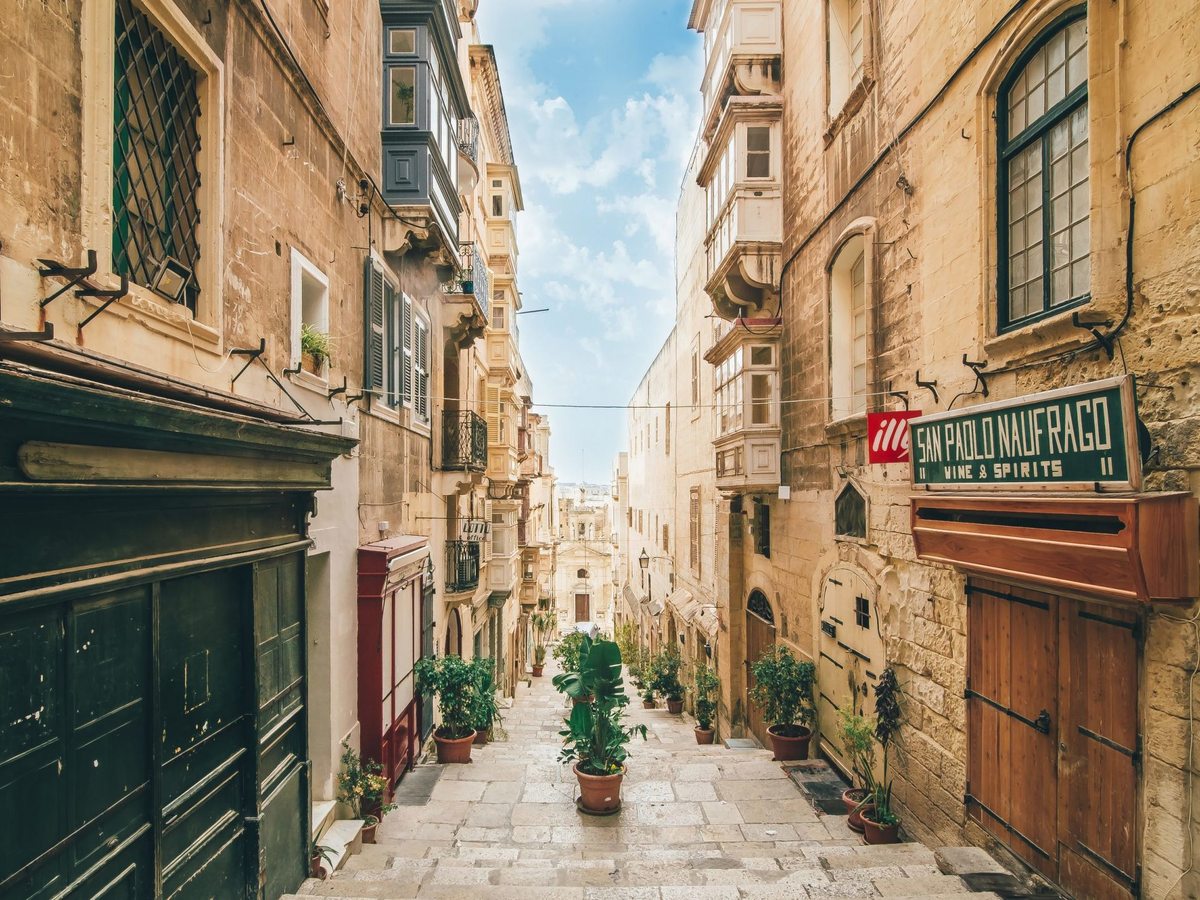 A narrow street of honey-coloured limestone buildings with enclosed wooden balconies, stepping down between potted plants towards a church front at the far end.