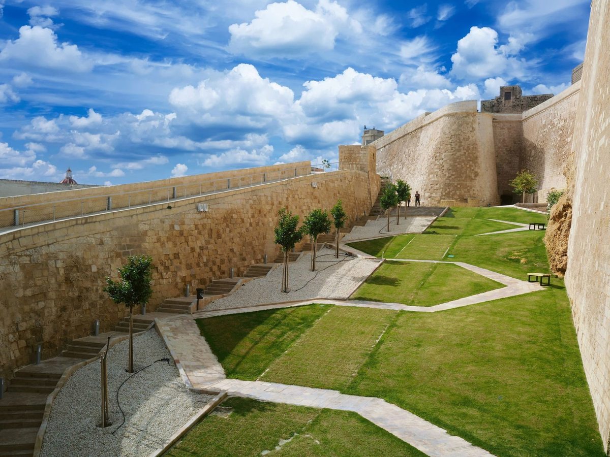 High stone bastion walls above a landscaped ditch of lawns, gravel paths and young citrus trees, under a bright sky with white cloud.